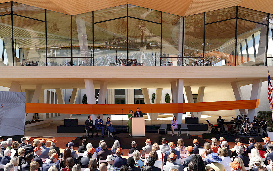Photo of Harriet and Warren Stephens at a podium on a stage in front of a large orange ribbon wrapped around the courtyard entrance to AMFA on opening day.