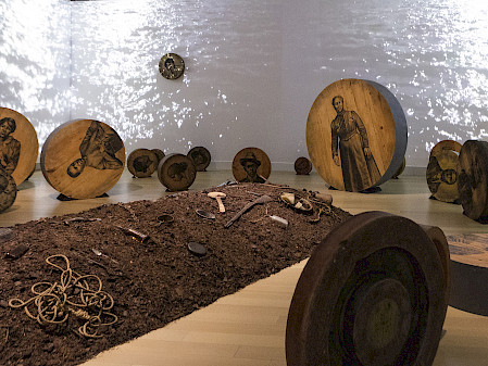 Photo of an artwork installation featuring a mound of dirt on the floor covered in objects surrounded by charcoal drawings of people on large round pieces of wood with a video of water projected on the walls behind them.