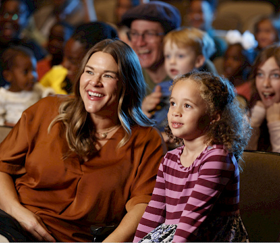 Photo of a mother and young daughter sitting in a theater surrounded by other parents and children watching a performance.