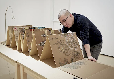 Photo of Sun Xun wearing a black shirt, gray pants, and black glasses leaning over a table holding his paintings on one long, folded stretch of board.
