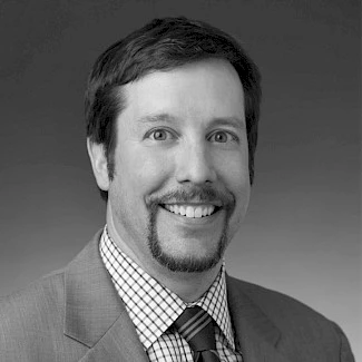 Square black and white photo of a white man with a goatee smiling and wearing a suit, patterned shirt, and striped tie.